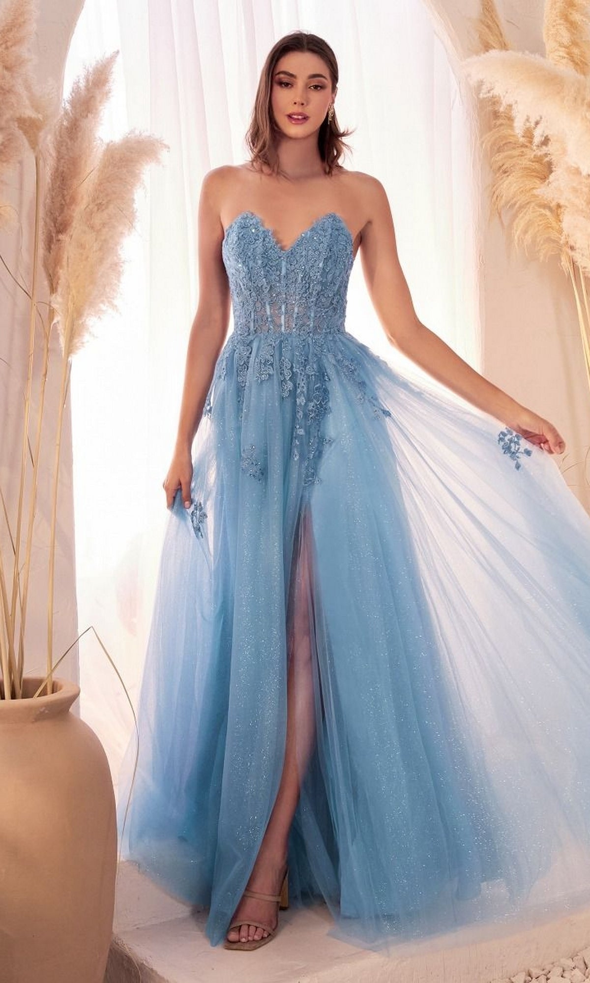 A woman poses indoors beside pampas grass, wearing the Strapless Long Glitter-Tulle Prom Ball Gown C148—a light blue, floor-length dress with floral appliqué, a front slit, and a strapless neckline.